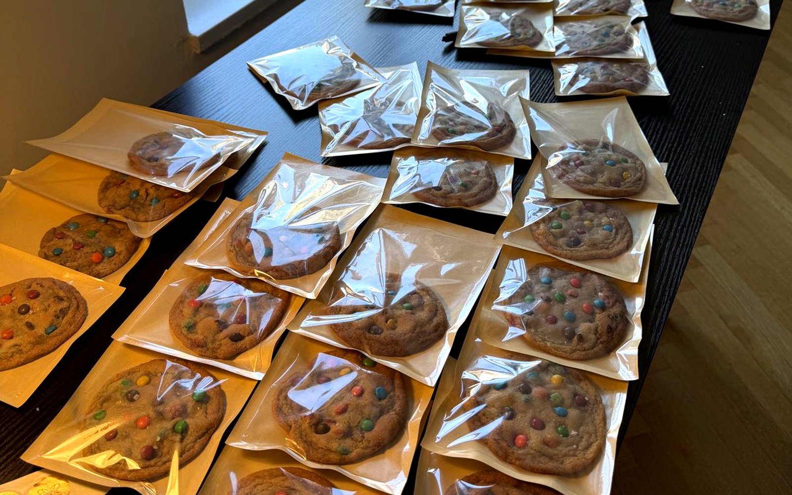 Dozens of cookies sealed in kraft and glassine, lined across a dark table in low afternoon light